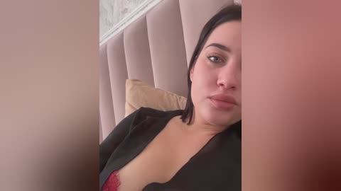 Video of a woman with light skin, dark hair, and full lips, wearing a black jacket and red bra, leaning against a beige headboard.