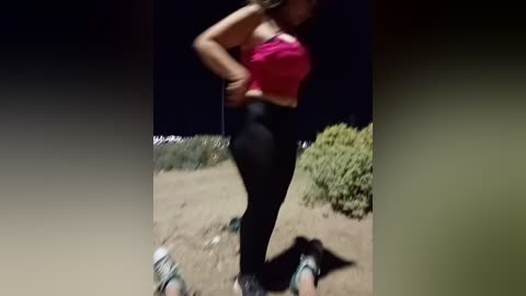 Video of a woman in a pink sports bra and black leggings, running outdoors at night, with blurred background showing grass and bushes.