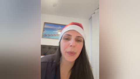 A video of a young woman with long dark hair, wearing a red Santa hat, a dark coat, and making a kissy face in a dimly lit room with a framed beach scene on the wall.