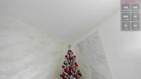 Video of a minimalist, white-walled room with a small, decorated Christmas tree adorned with red and gold ornaments and a star topper, positioned in the corner. The room's ceiling is angular, creating a modern, geometric look.