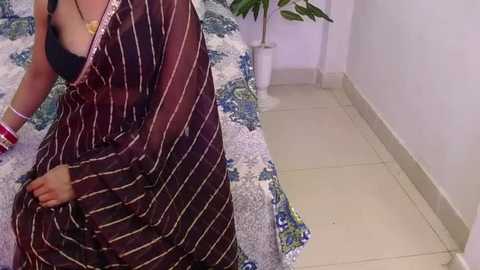 Video of a woman in a brown and white striped sari, sitting on a bed with a floral patterned bedspread, wearing a black bra.