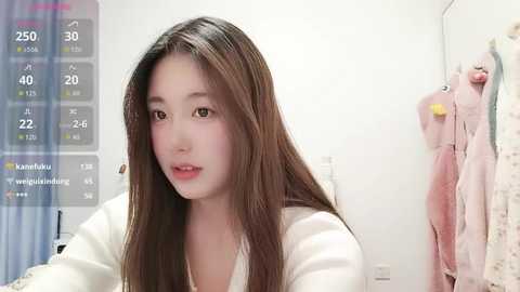 A video of an East Asian woman with long brown hair and fair skin, wearing a white robe, standing in a bathroom with a digital scale on the left and a towel rack on the right.
