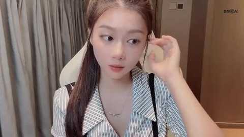 Video of an Asian woman with long, straight brown hair, wearing a striped shirt, adjusting her hair in a modern room with beige curtains and a white chair.