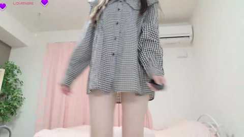 Video of a woman in a checkered coat, standing on a bed in a bright room with pink curtains and a green plant.