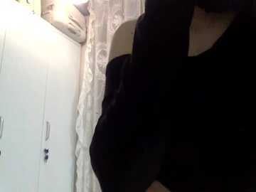 A video shows a person in a black shirt leaning against a white cabinet with a patterned curtain in the background. The image is dimly lit, creating a shadowy effect.