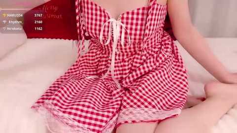 A video of a light-skinned woman wearing a red and white gingham nightgown with lace trim, seated on a white bed.