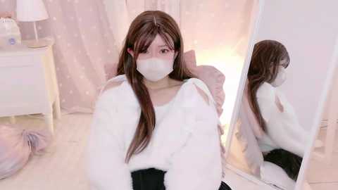 Video of an Asian woman with long brown hair, wearing a white mask, fluffy white sweater, and black pants, kneeling in front of a mirror in a softly lit, pastel-colored room.