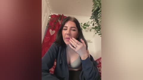 A video of a young woman with medium skin tone, long dark hair, and a medium-sized bust, wearing a light blue bra and a dark cardigan. She has her hand to her mouth, looking playful. Background includes a red heart-patterned curtain and green plants.