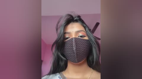 Video of a young woman with medium brown skin and long black hair, wearing a black face mask with a geometric pattern, a grey top, and a gold necklace. Background is a pink room with a ceiling fan.