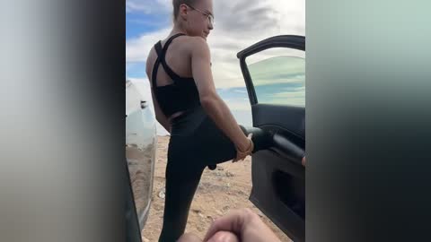 A video of a fit, blonde woman with glasses in a black sports bra and leggings, stretching in a car door. Desert landscape with white buildings in the background.