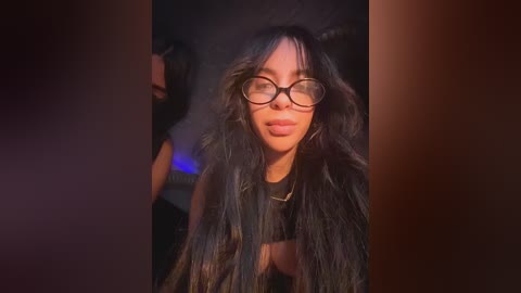 A video of a woman with long, wavy black hair, wearing black-framed glasses and a black outfit, standing in a dimly lit room with blurred background.
