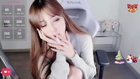 Video of a young Asian woman with long brown hair, wearing a beige sweater, sitting in a gaming chair, eyes closed, mouth open, hands on her cheeks. Background features a monitor with health metrics and a white desk with decorations.