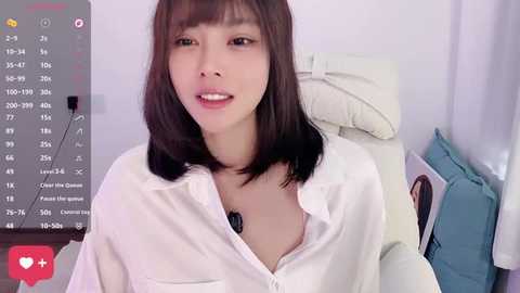 A video of an Asian woman with straight black hair, wearing a white button-up shirt, sitting on a white couch, with a microphone in front of her.