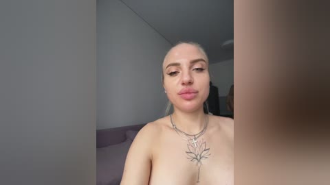 A video of a topless woman with fair skin and blonde hair, wearing a floral necklace, sitting indoors. The background includes a purple couch and a plain white wall.