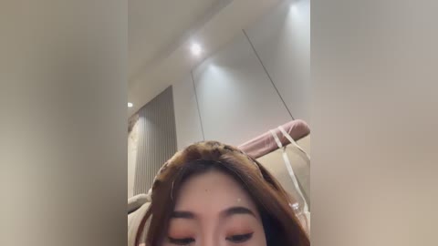 A video of a woman with long, light brown hair, wearing a beige robe, standing in a dimly lit room with white walls and a pink towel hanging on a door.