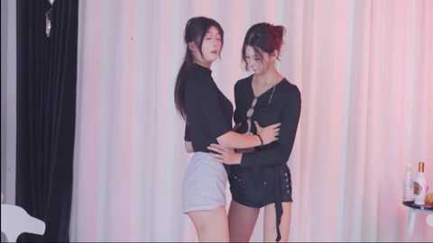 Video of two East Asian women with long dark hair, one in a black top and high-waisted shorts, the other in a black dress, standing close, arms crossed, against a white and pink background.