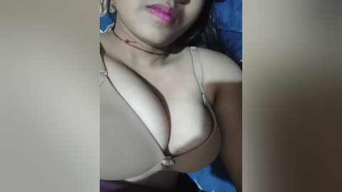 Media: A close-up video of a South Asian woman with medium skin tone, wearing a revealing beige bikini top that accentuates her large breasts. Her lips are painted pink, and she's lying on a blue blanket.