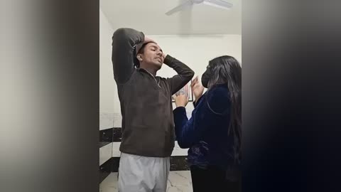 Video of a man in a black jacket and white pants stretching, while a woman in a navy blue dress and mask watches from the side in a dimly lit room.