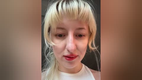 Video of a fair-skinned, blonde woman with bangs, wearing a white tank top, standing against a blurred, dark background. She has a neutral expression.
