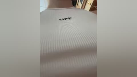 Media: A video of a person wearing a white, ribbed, long-sleeved shirt with a small, black, \"CF\" logo centered on the chest. The background features a blurred, indoor setting with wooden furniture and a beige wall.