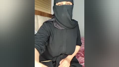 Video of a light-skinned woman in a black hijab and long-sleeved dress, sitting on a bed with a pink blanket, in a dimly lit room.