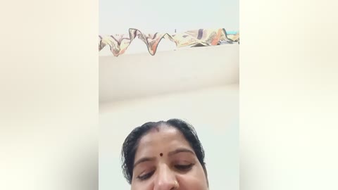 Video of a woman with dark hair and fair skin, wearing a saree, standing indoors. Her eyes are closed, and she has a bindi on her forehead. A colorful, patterned cloth hangs on a wall behind her.