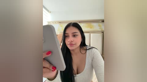 Video of a young woman with long black hair and fair skin, wearing a low-cut gray top, taking a selfie with a silver smartphone in a bathroom with a floral-patterned shower curtain and beige walls.