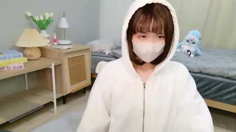 A video of a young Asian woman in a white hoodie with a white mask, sitting in a modestly furnished room with a bed and nightstand.
