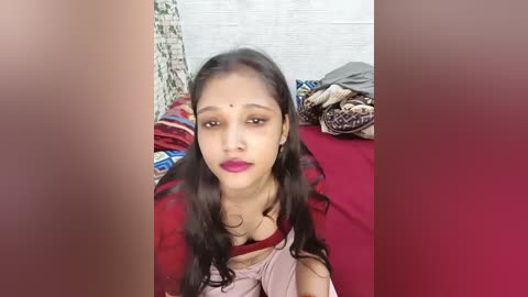 A video of a young South Asian woman with long black hair, wearing a red sari with a pink blouse, sitting on a bed with a red sheet, surrounded by colorful patterned blankets and a wall with a white brick texture.
