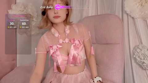 A video of a young Asian woman with fair skin and blonde hair in a pink, frilly lingerie outfit, sitting on a plush pink chair, surrounded by soft, pastel decor.