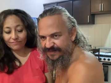 A video of a middle-aged woman with long dark hair and a man with long gray hair and a beard, both smiling, standing in a modern kitchen with wooden cabinets and stainless steel appliances.