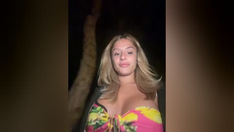 Media: Video of a young, light-skinned woman with shoulder-length blonde hair, wearing a colorful, strapless top, standing indoors against a dimly lit, dark background.