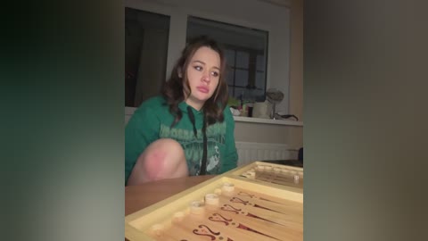 A video of a young woman with long brown hair, wearing a green hoodie, sitting at a wooden chessboard, indoors with dim lighting.