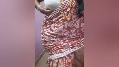 Video of a pregnant woman with dark skin, wearing a red floral-patterned sarong and yellow beaded necklace, holding a white pillow, against a lavender wall.