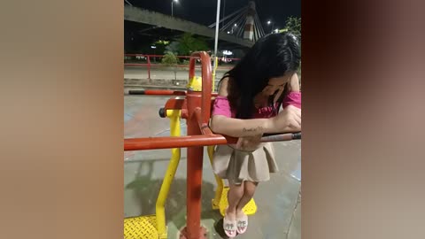 Video of a young Asian woman with long black hair, wearing a pink shirt and beige skirt, sitting on a red playground slide, looking sad, at night under a bridge.