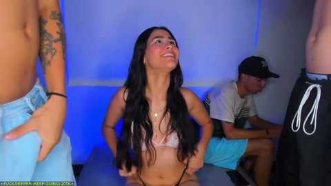 Media: Video of a young woman with long black hair, wearing a pink lace bra, sitting on a table, surrounded by shirtless men in shorts, and a man in a baseball cap, in a dimly lit room.