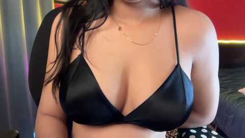 Video of a woman with medium skin tone, long dark hair, wearing a black satin bralette that accentuates her ample cleavage. She has a thin gold necklace and a leopard-print skirt in the background.