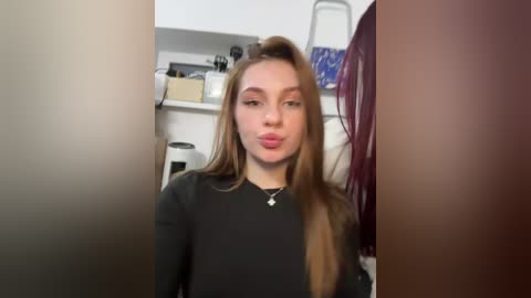 Media: A video of a young Caucasian woman with long, straight, light brown hair, wearing a black long-sleeved top, pouting with her lips, in a cluttered kitchen with white cabinets and a purple towel.