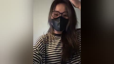 A video of a young Asian woman with long brown hair, wearing black glasses, a black face mask, and a black-and-white striped shirt. She stands against a plain, light-colored wall, with her right hand touching her head.