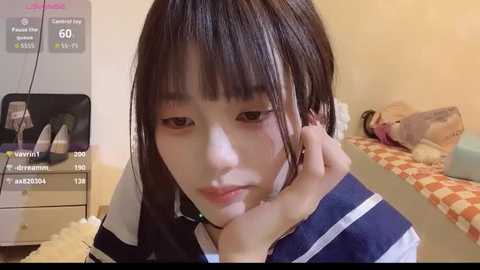Video of a young Asian girl with straight, shoulder-length brown hair, wearing a navy sailor-style school uniform, resting her head on her hand, indoors with a white wall and a checkered cushioned bench in the background.