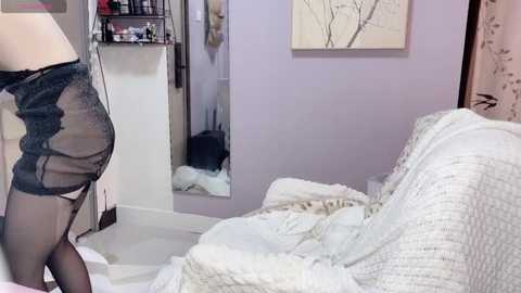 Media: Video of a woman in black lingerie, garter belt, and stockings, bending over in a bedroom with white sheets, a mirror, and light purple walls.