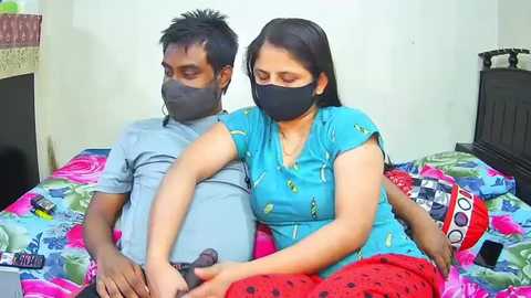 Video of an Indian couple wearing face masks, sitting on a colorful bedspread in a simple bedroom with a nightstand and wall decor.