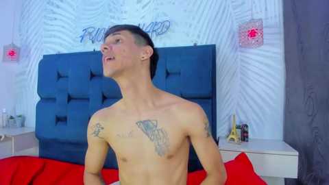 Video of a shirtless, slim, fair-skinned young man with short dark hair, tattoos on his chest, and a tattooed arm, standing in a bedroom with white walls and a blue tufted headboard.