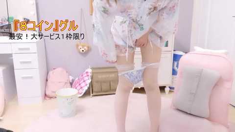 A video of a slender, fair-skinned woman in a pastel floral kimono, partially pulling down blue and white patterned panties in a pink-themed, cluttered bedroom.