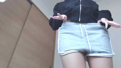 Media: Video of a person from the waist down, wearing a black jacket and light blue shorts with white trim. They stand in front of a wooden wardrobe, with hands gesturing.
