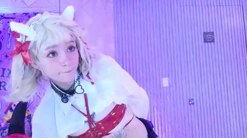 Video of a young woman with pale skin and blonde hair styled in a bob, wearing a white and red anime-inspired maid outfit with cat ears, against a purple-striped background.