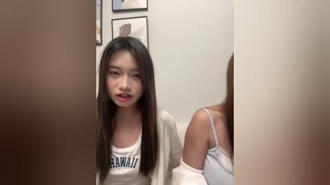 Video of a young East Asian woman with long dark hair, wearing a white \"Baylor University\" tank top and white cardigan, standing in a beige-walled room with framed art.