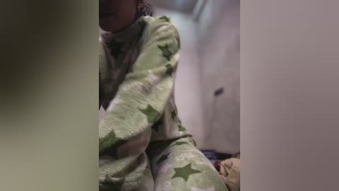 Media: Video of a person in green pajamas with star patterns, sitting on a bed in a dimly lit room, partially obscured by a dark figure in the foreground.