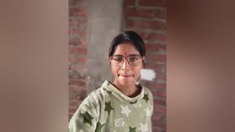 Media: Video of a woman with medium brown skin, short black hair, and glasses, wearing a green hoodie with white star pattern, standing against a brick wall.