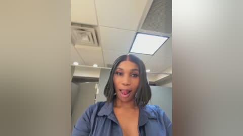 A video of a young woman with medium brown skin and straight black hair, wearing a blue denim jacket and sticking out her tongue. She stands in a modern, brightly lit room with white walls and a recessed ceiling light.